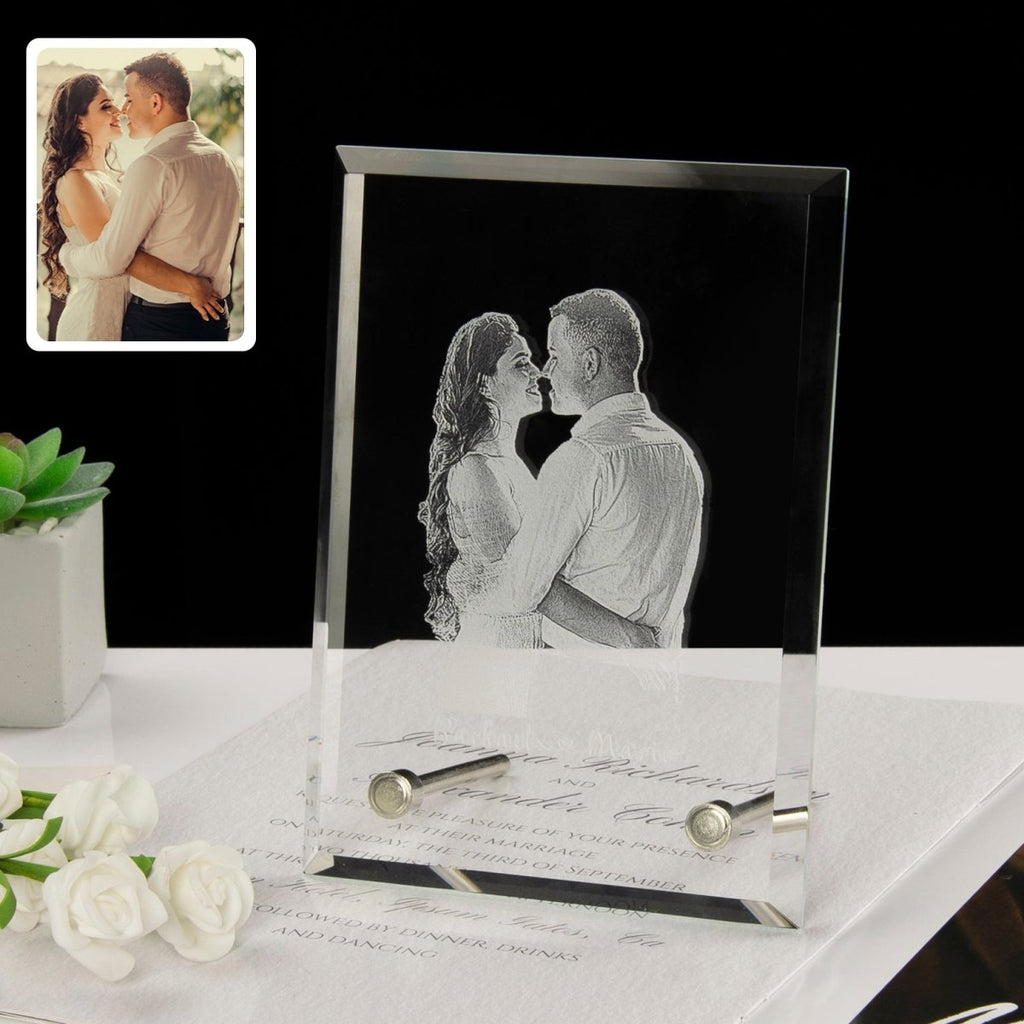Glass photo frame with engraved couple's photo on a desk with flowers and stationery.
