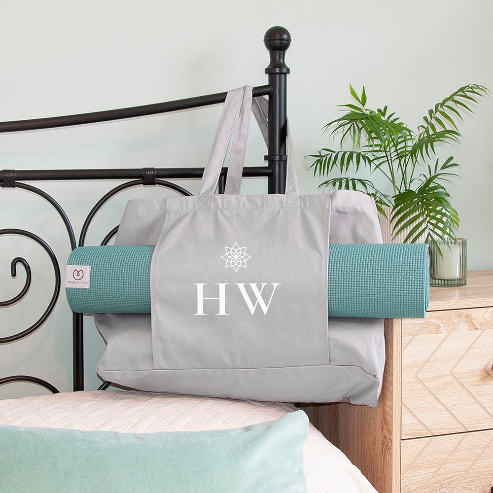 Gray tote bag with 'HW' initials on a bed with yoga mats and a plant in the background