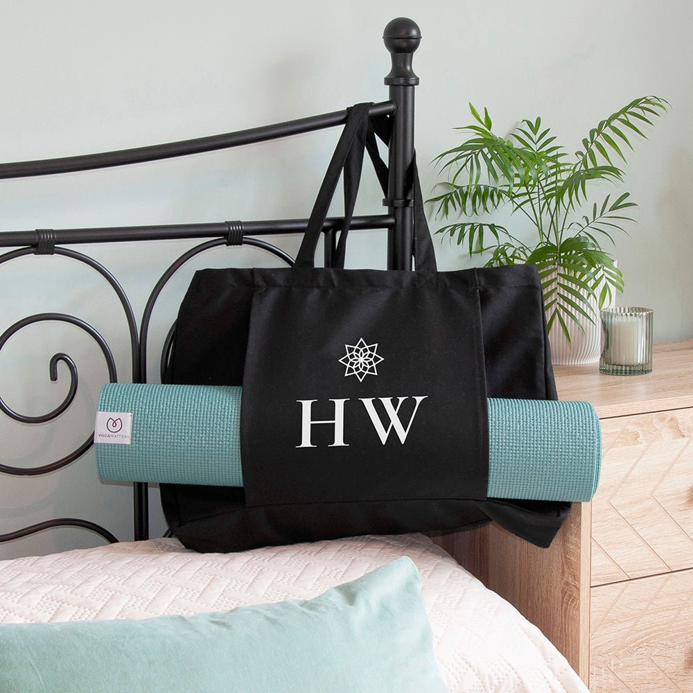 Black tote bag with initials 'HW' on a bed with a green yoga mat underneath.