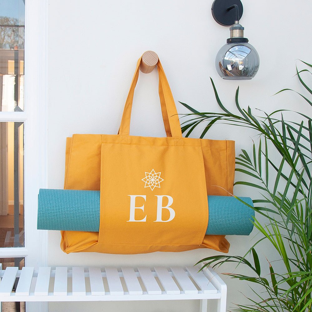 Yellow tote bag with 'EB' monogram and blue yoga mat hanging on a hook against a white wall.