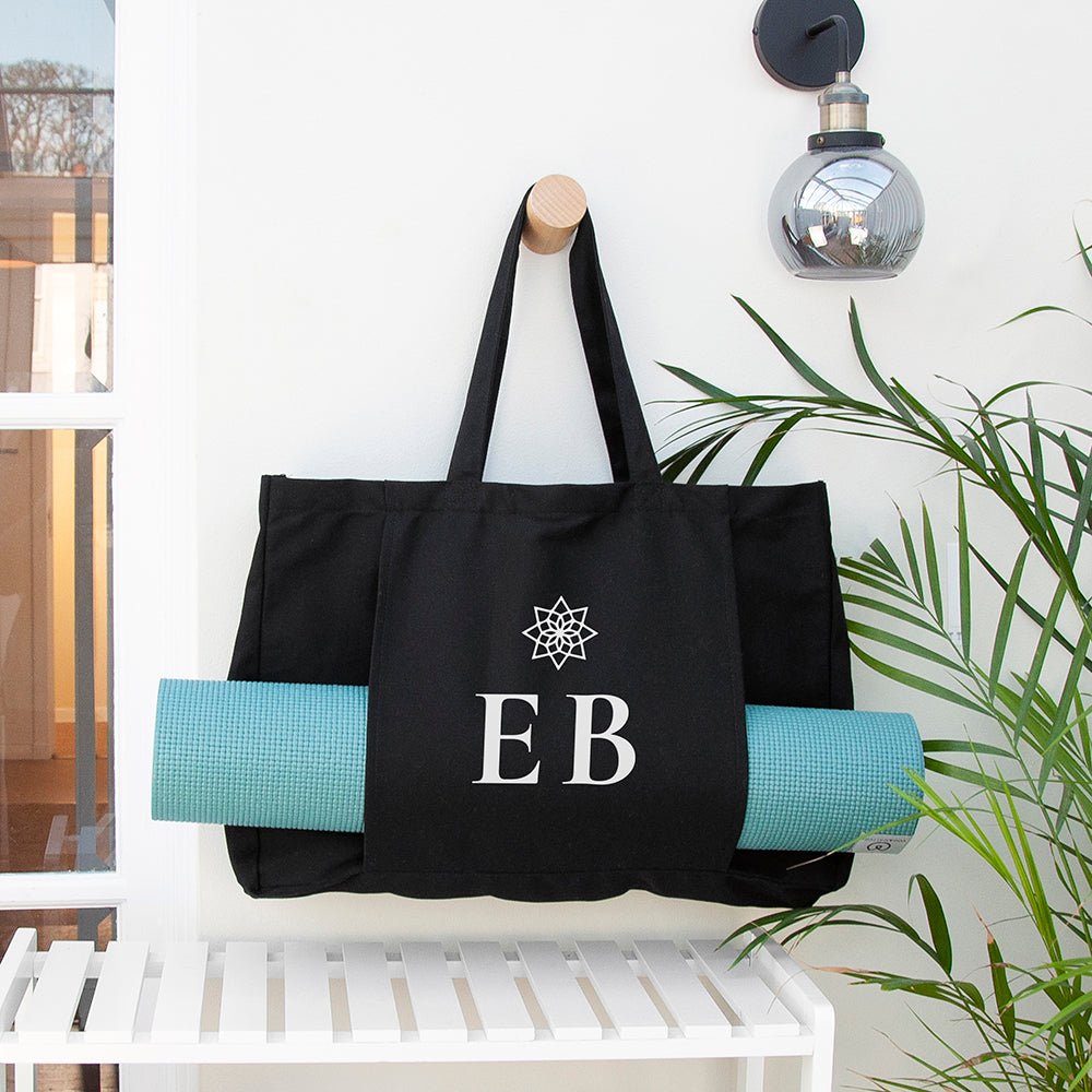 Black tote bag with 'EB' initials and a blue yoga mat on a white bench.