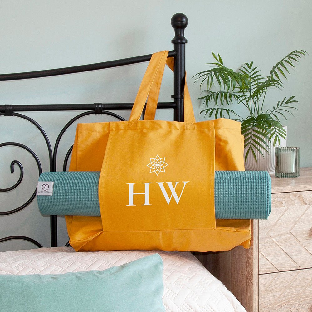 Yellow tote bag with 'HW' initials on a bed with a yoga mat underneath, plant in the background
