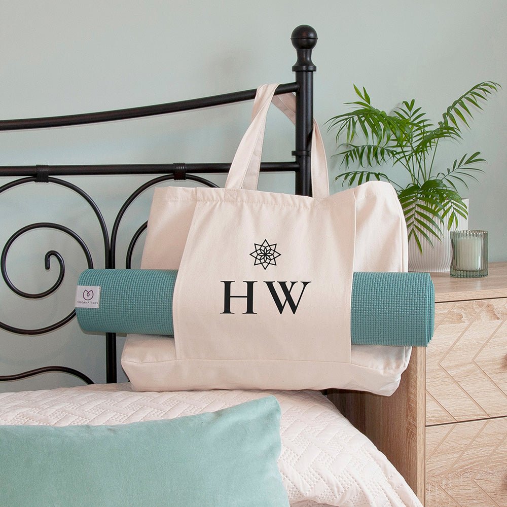 White tote bag with 'HW' logo on a bed with rolled-up yoga mat and pillow