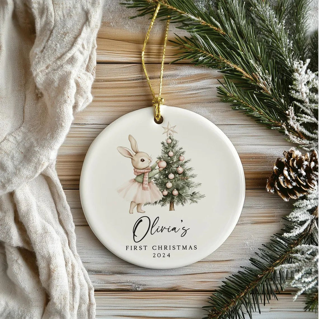 Decorative Christmas ornament with a rabbit and tree design, featuring 'Olivia's First Christmas 2024' text, on a wooden surface with pine branches and pinecones.