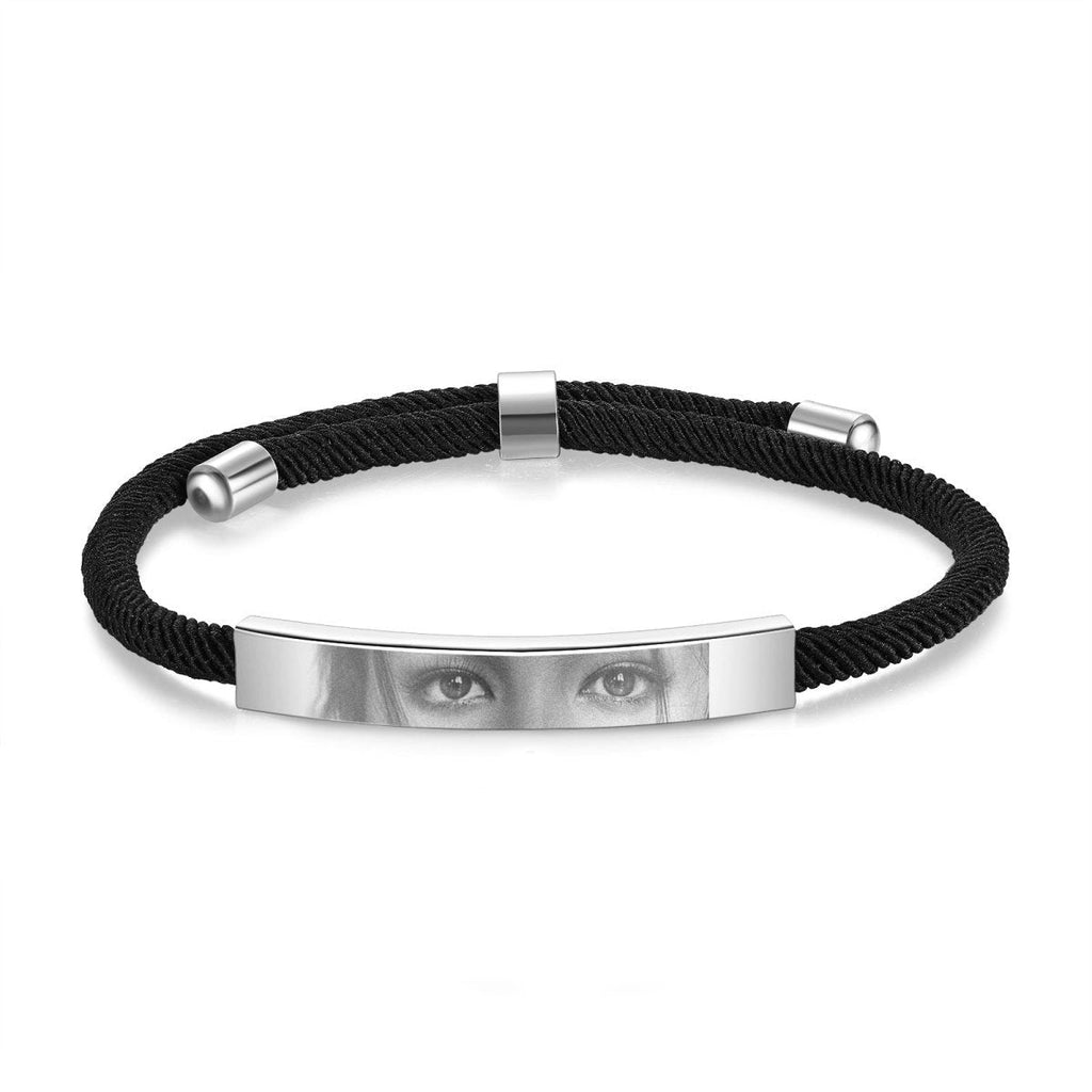 A black braided bracelet with a silver metal bar featuring a printed image of a pair of eyes.