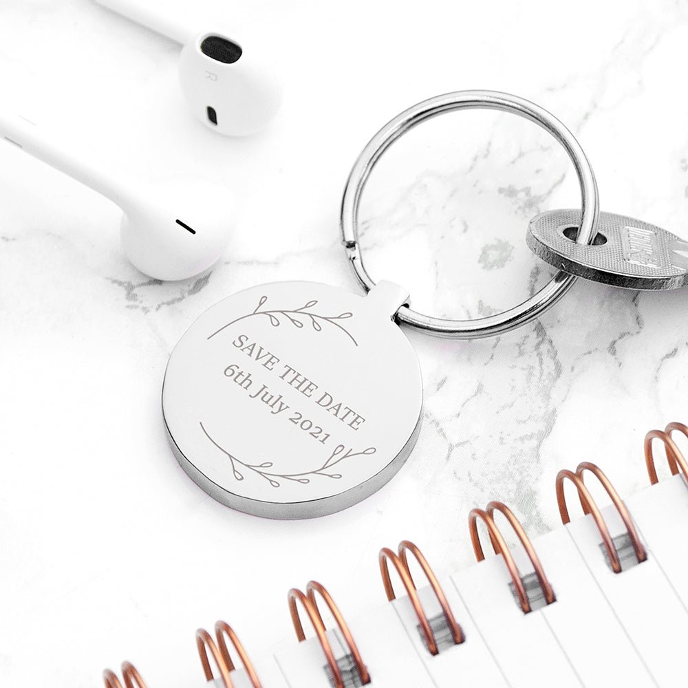 Silver keychain with 'Save the Date' engraving on a marble surface with white earbuds and notebooks.