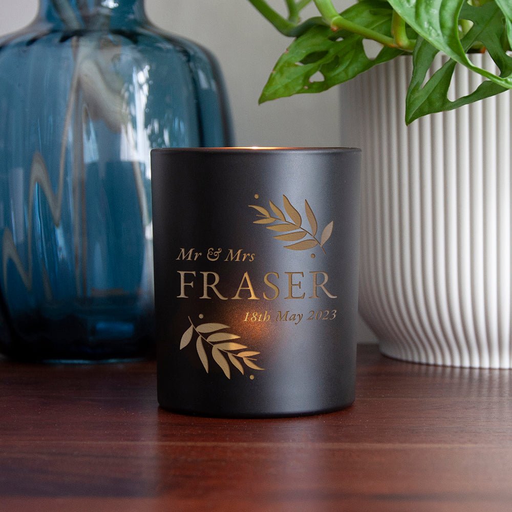 Black candle with 'Mr & Mrs Fraser' engraving on a wooden surface with decorative vases.